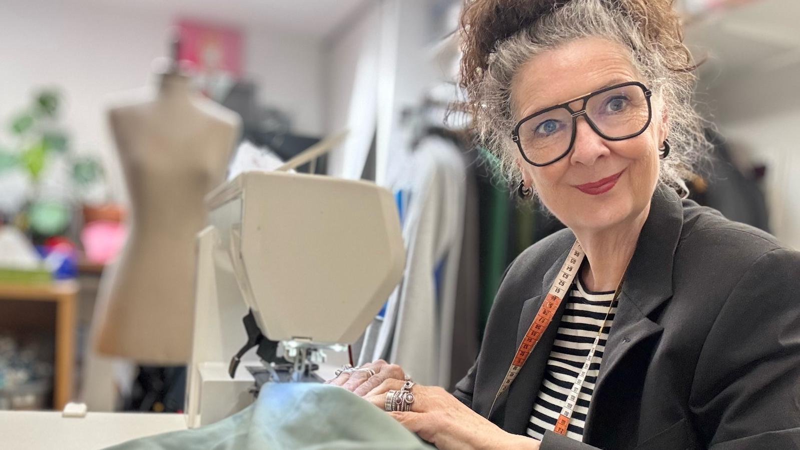 Lachende modeontwerpster Claudia Träumer van CLAU-D Handmade Design, met een meetlint om haar nek en grote zwarte bril, aan het werk achter een naaimachine in haar atelier in Arnhem. Op de achtergrond een paspop en kledingrekken.